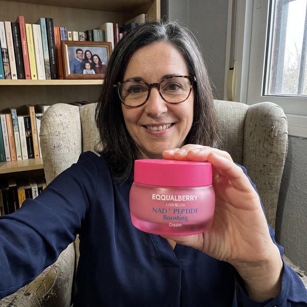 Mujer de unos 52 años sentada en un sillón de su salón, sonriendo, con el tarro rosa de EQQUALBERRY en la mano. Detrás, una estantería con libros y una foto de familia.