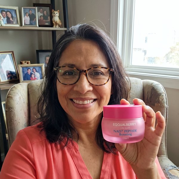 Mujer de unos 52 años sentada en un sillón de su salón, sonriendo, con el frasco rosa de EQQUALBERRY en la mano. Detrás, una estantería con libros y una foto de familia.