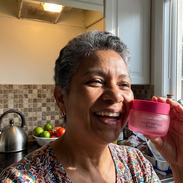 Mujer de unos 58 años en la cocina de su casa, con el pelo corto entrecano, sujetando el frasco rosa de EQQUALBERRY junto a la cara. Al fondo, azulejo, un hervidor y un frutero.