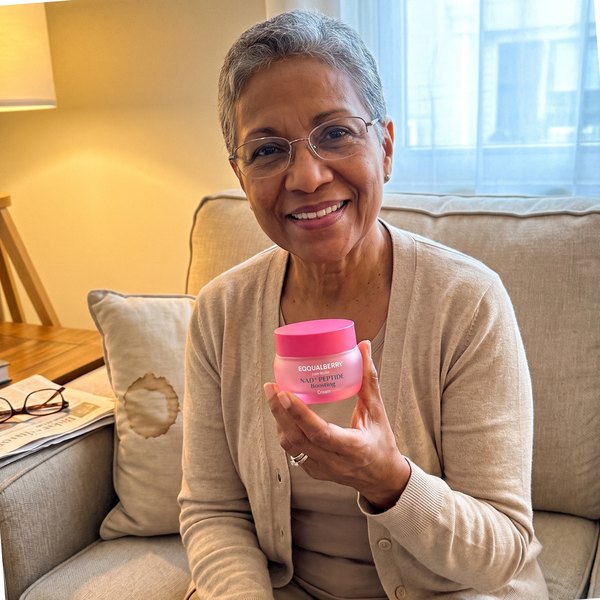 Mujer de unos 63 años sentada en un sofá beige, con gafas y una rebeca, sosteniendo el frasco rosa de EQQUALBERRY. En la mesita, unas gafas de leer y un periódico.